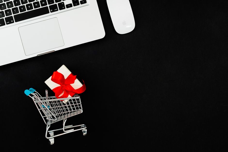 Minimalist image of a shopping cart with a gift on a black background beside a laptop, ideal for ecommerce themes.